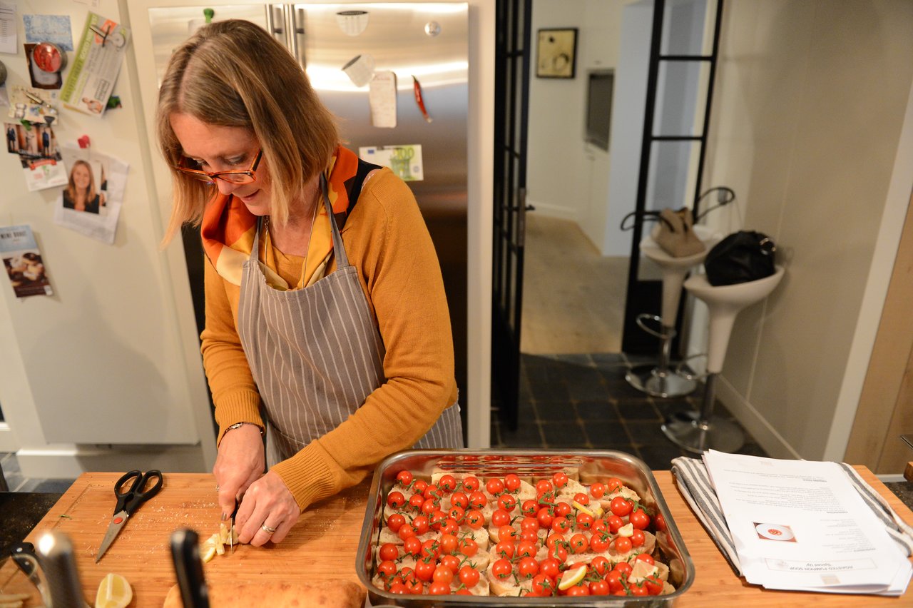 A woman wearing an apron chops ingredients on a wooden board next to a tray of cherry tomatoes and bread.