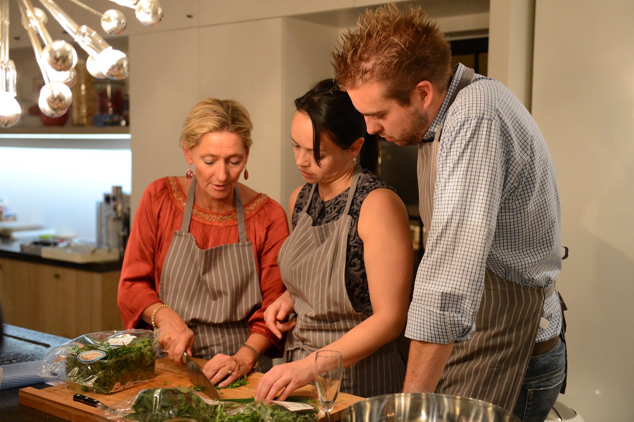 A woman in an apron demonstrates chopping herbs while two others watch closely during a cooking class.