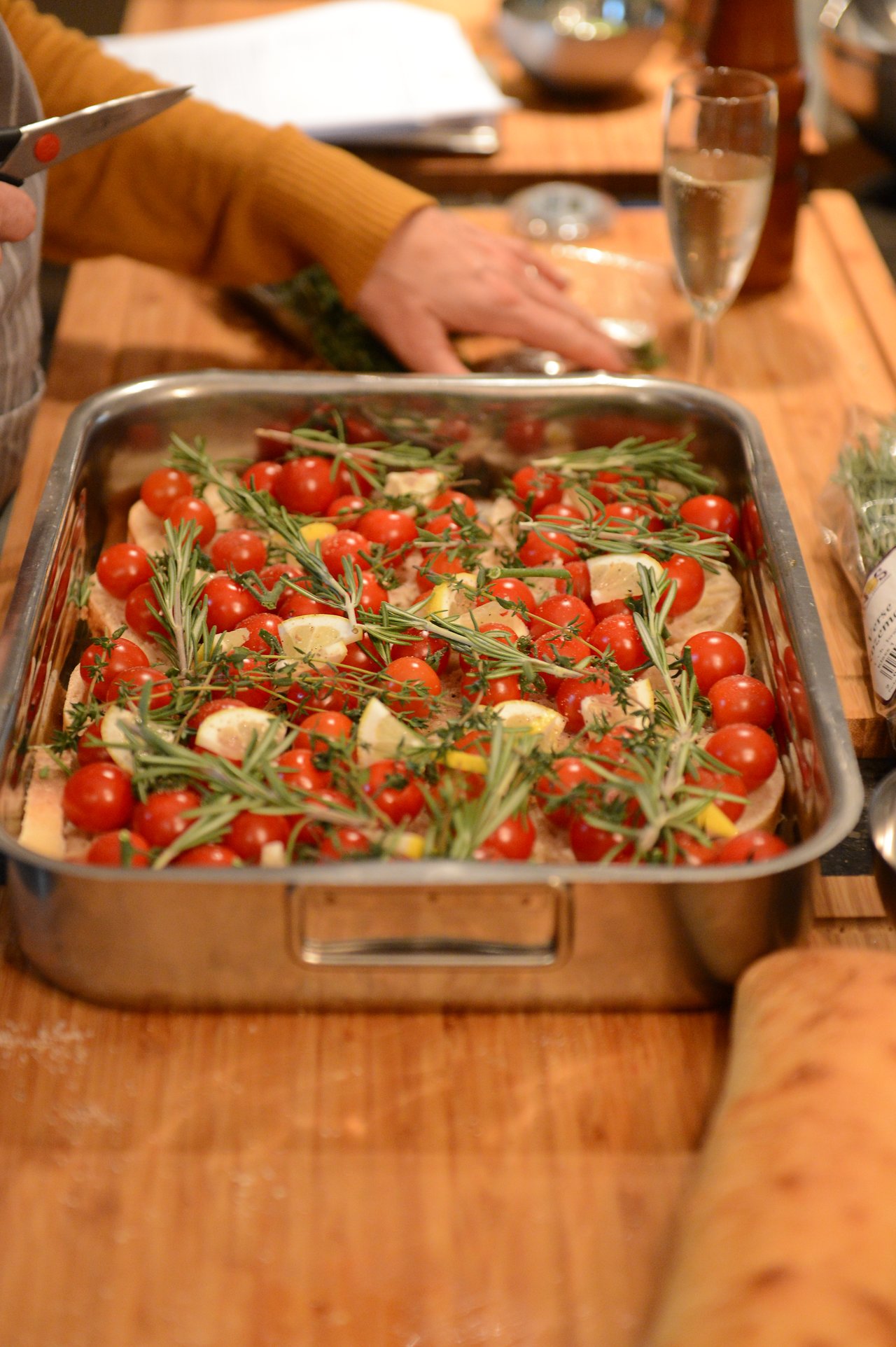 A person prepares a baking dish with chicken, cherry tomatoes, lemon slices, and rosemary during a cooking class.