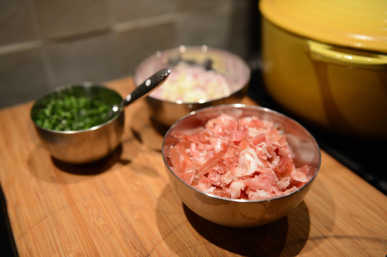 Metal bowls with chopped ingredients, including meat, onions, and herbs, are placed on a wooden cutting board.