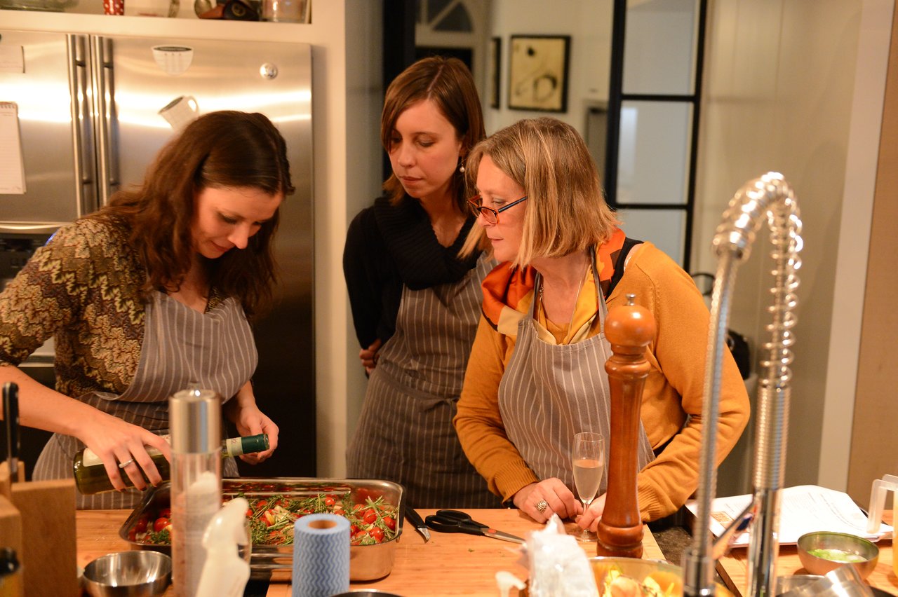 Three women in aprons stand around a kitchen counter, watching as one pours oil onto a tray of vegetables.