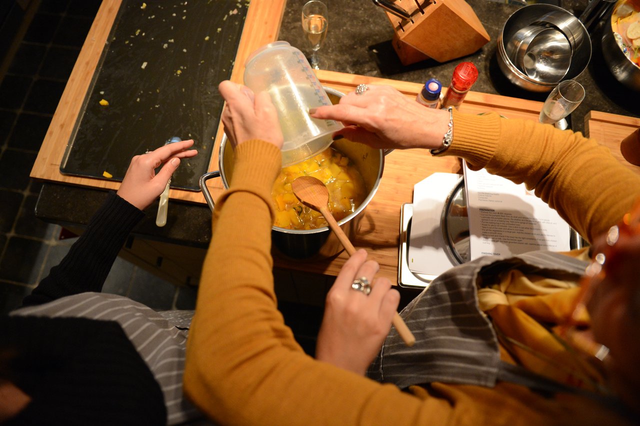 Two people in aprons prepare food, pouring ingredients into a pot and stirring during a cooking class.