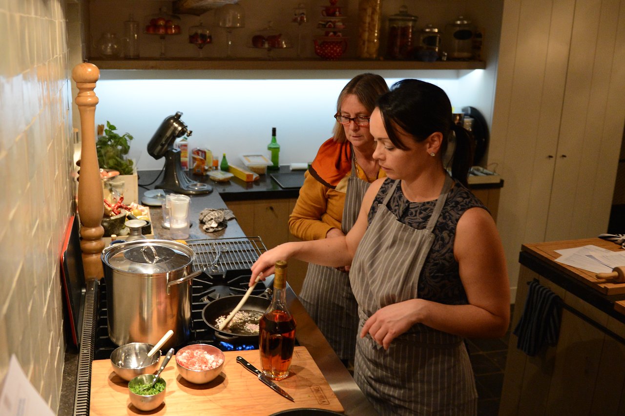 Two women in aprons cooking at a stove, one stirring a pan while the other observes.