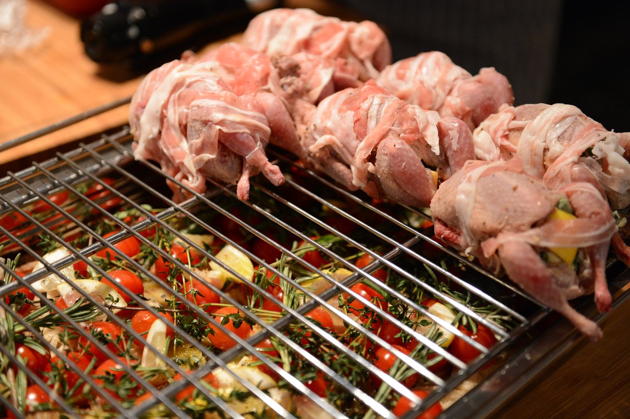 Raw, bacon-wrapped poultry on a grill above a tray of vegetables with herbs, prepared during a cooking class.