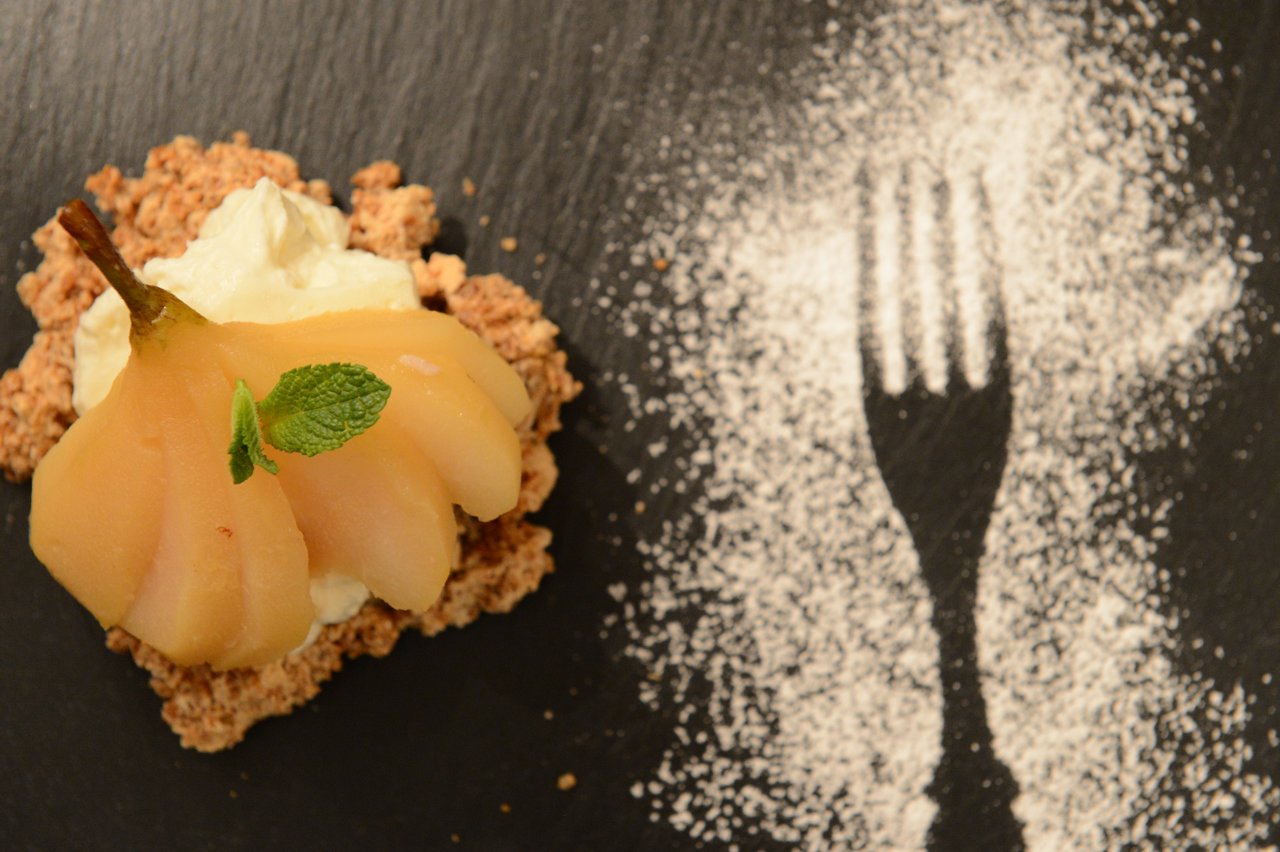 A plated dessert with poached pear, cream, and crumble, next to a fork silhouette made with powdered sugar.