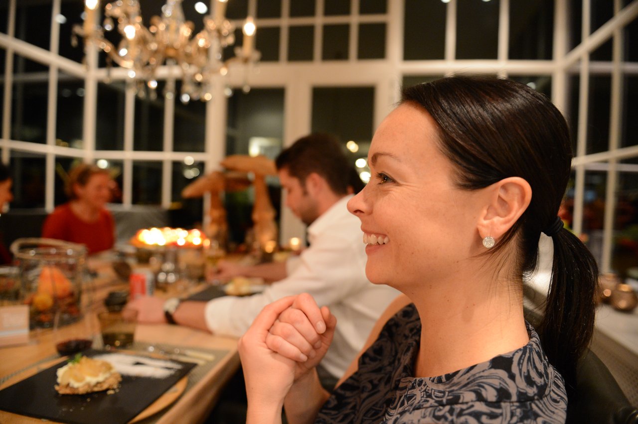 A smiling woman sits at a dinner table with others, enjoying a meal in a well-lit setting.
