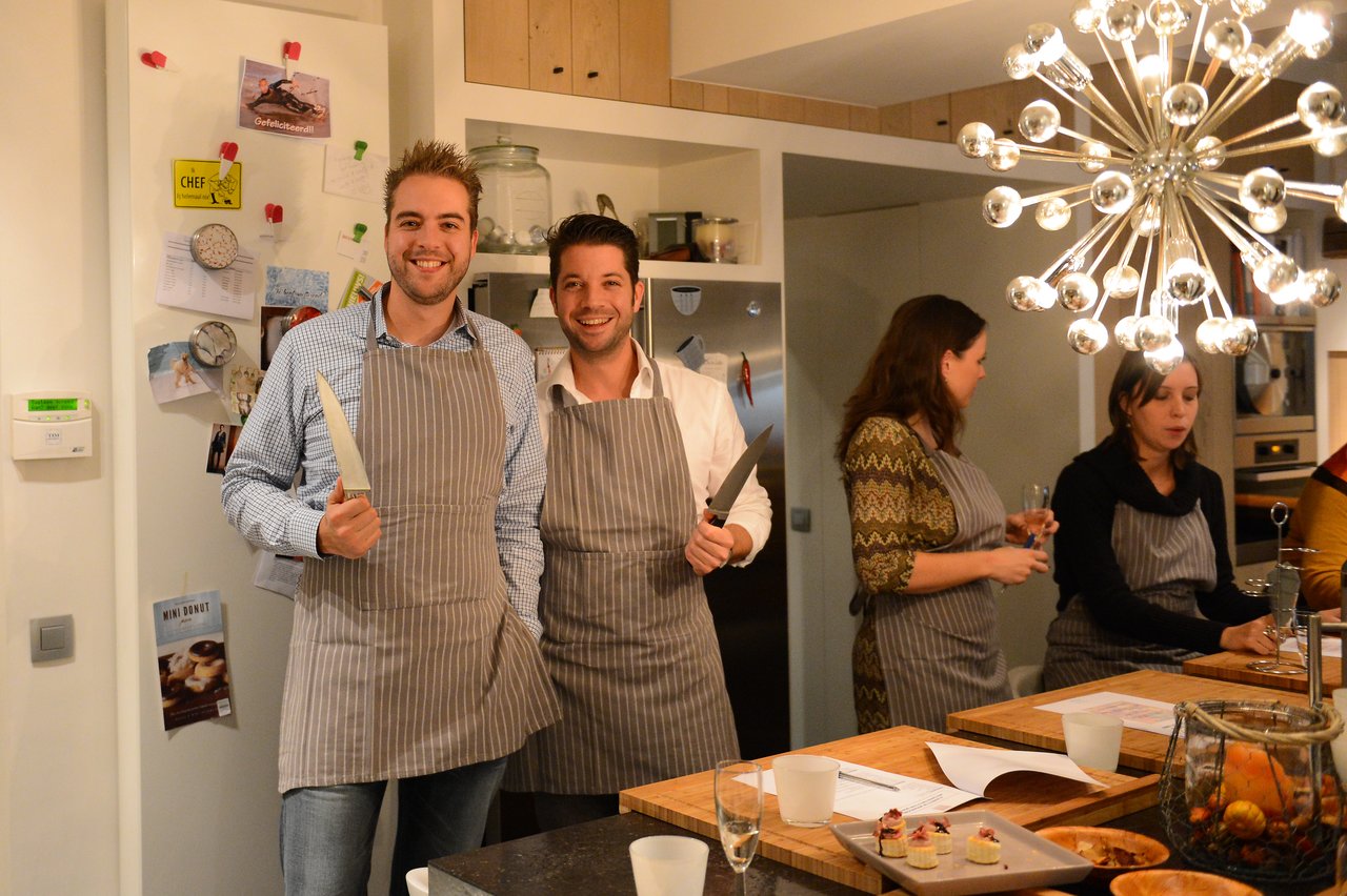 Two smiling men in aprons hold knives, posing for the camera during a cooking class.