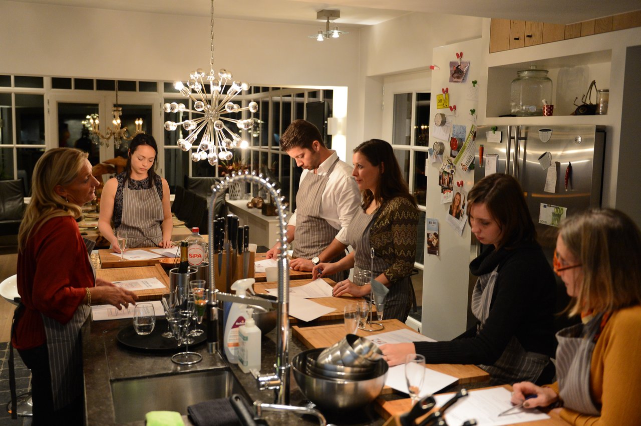 A group of people in aprons listen to an instructor while following a cooking lesson in a kitchen.