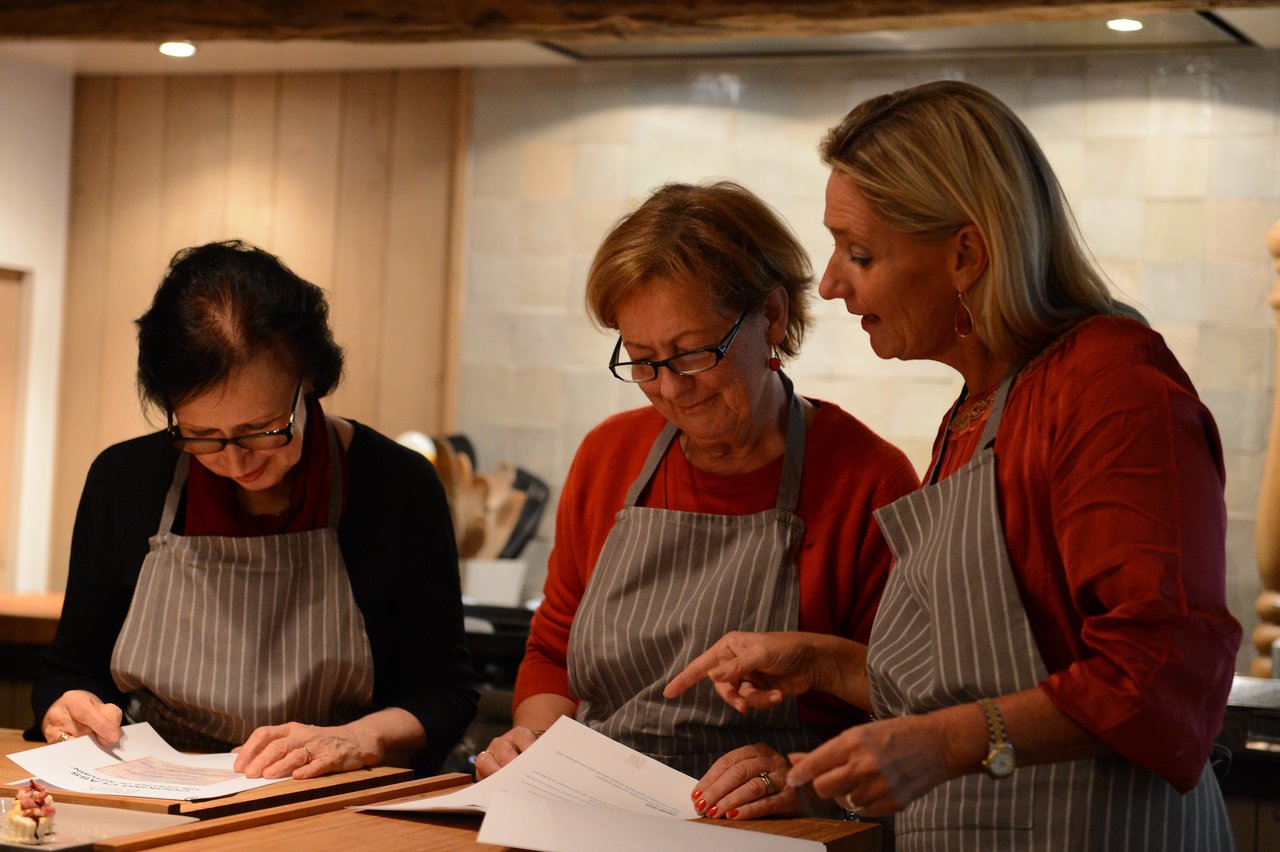 Three women in aprons review a recipe together during a cooking class, with one pointing at the paper.