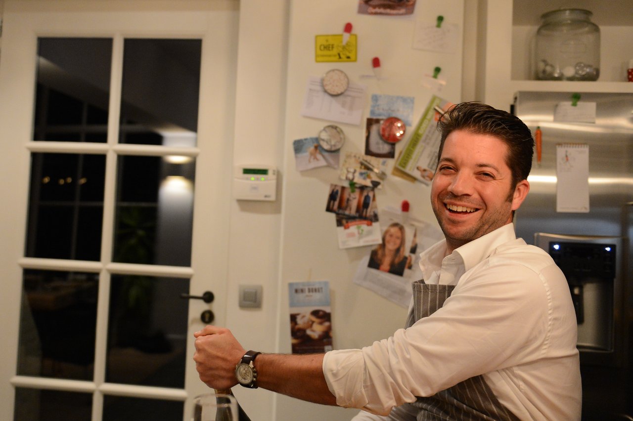 A smiling man in an apron sits at a table, holding a bottle in a cooking class.