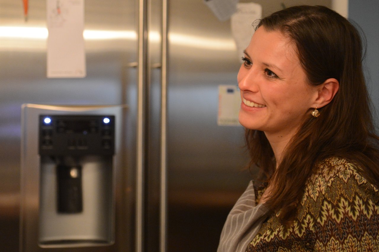 A woman wearing an apron smiles while standing in a kitchen near a refrigerator.