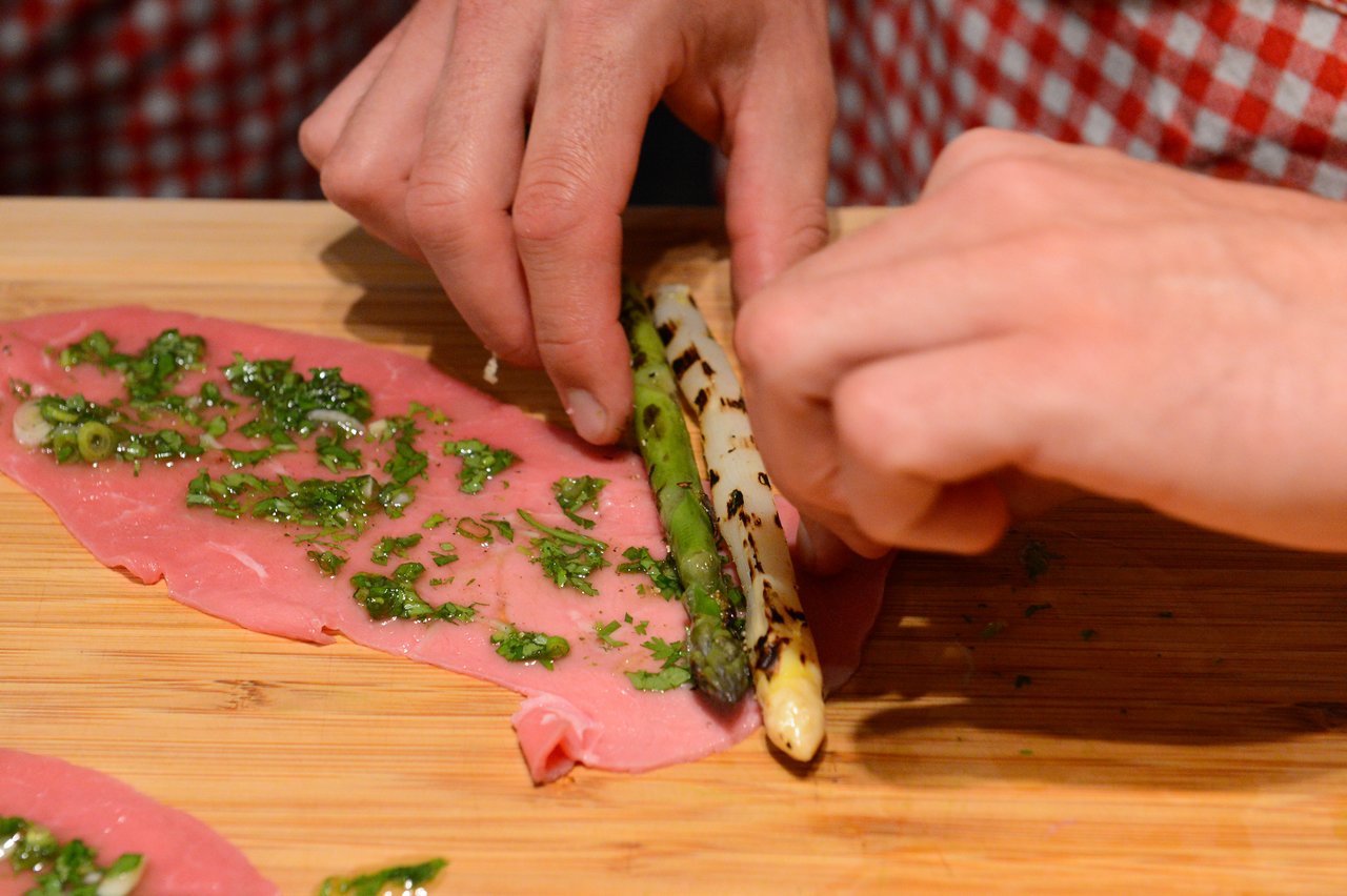 Hands rolling asparagus and herbs inside a thin slice of meat on a wooden cutting board.