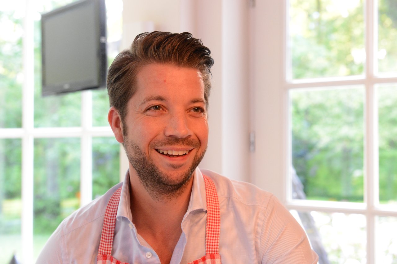 A smiling man wearing a checkered apron participates in a cooking class indoors with natural light from large windows.