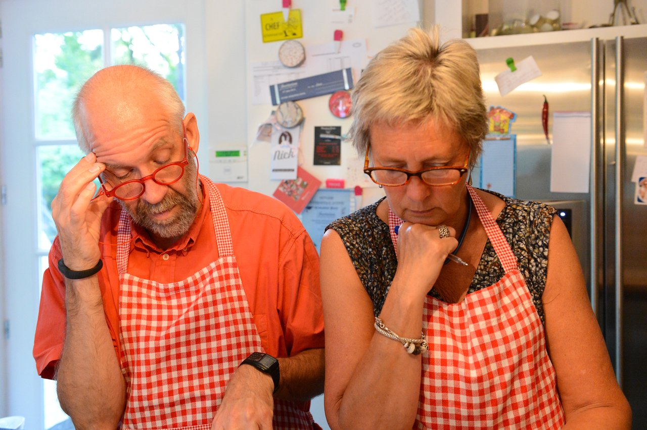 Two people wearing red checkered aprons and glasses focus intently on a cooking task in a kitchen.