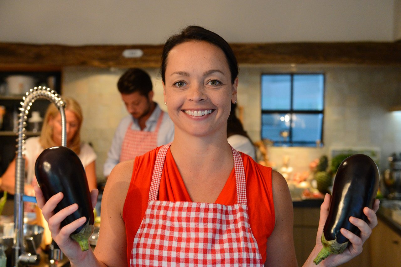 A woman in a red apron smiles while holding two eggplants in a cooking class.
