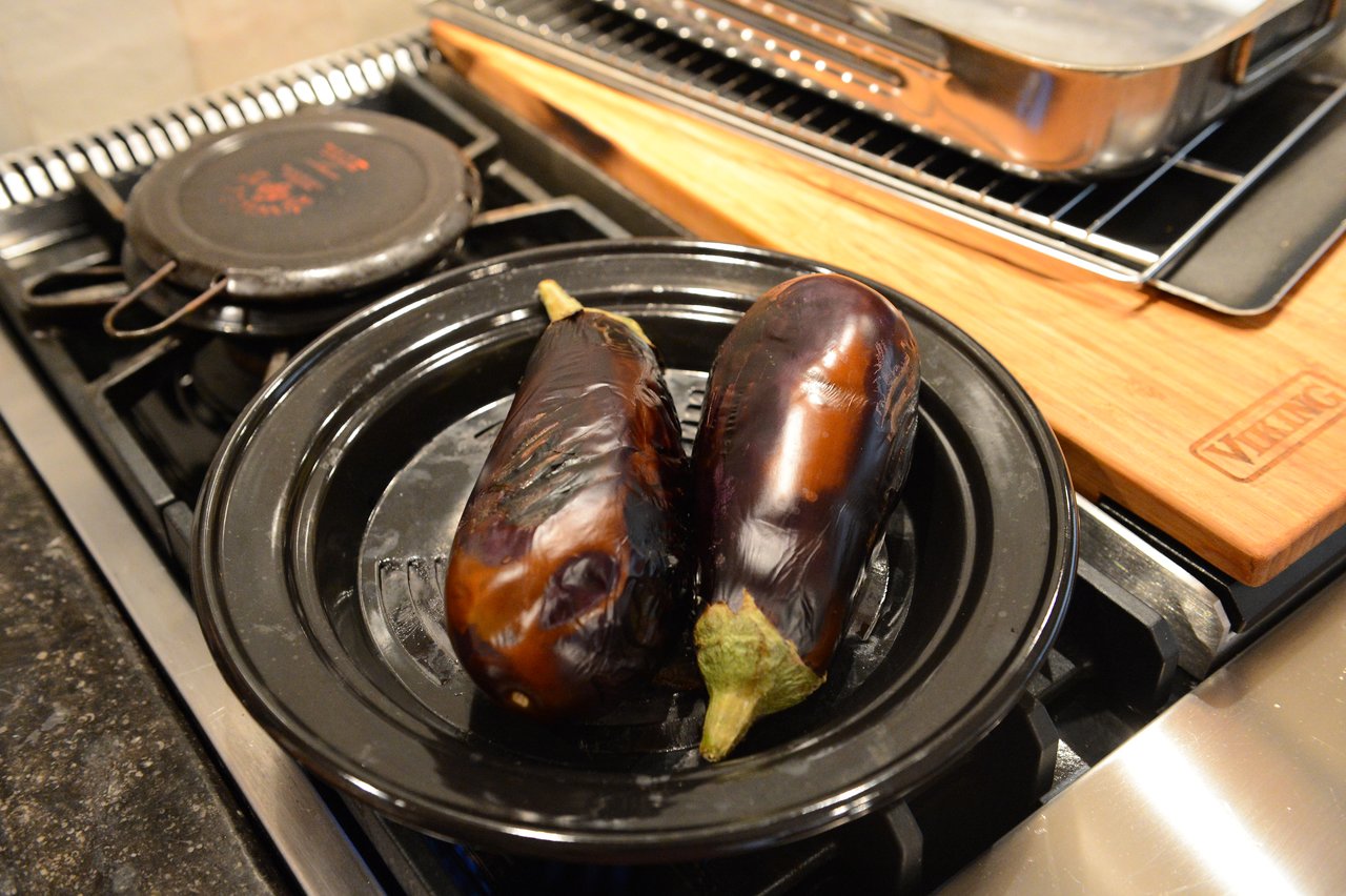 Two roasted eggplants rest on a black plate atop a stovetop during a cooking class.