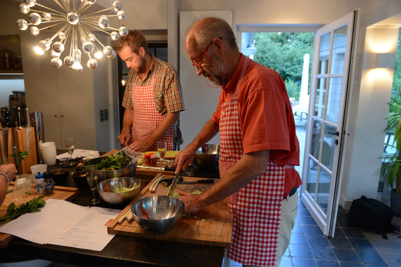 Two people wearing aprons chop ingredients on cutting boards during a cooking class in a well-lit kitchen.
