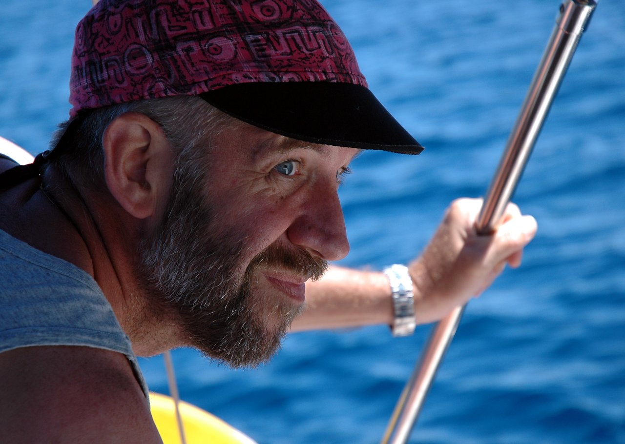 A bearded man in a red cap smiles while holding onto a metal railing near the water.