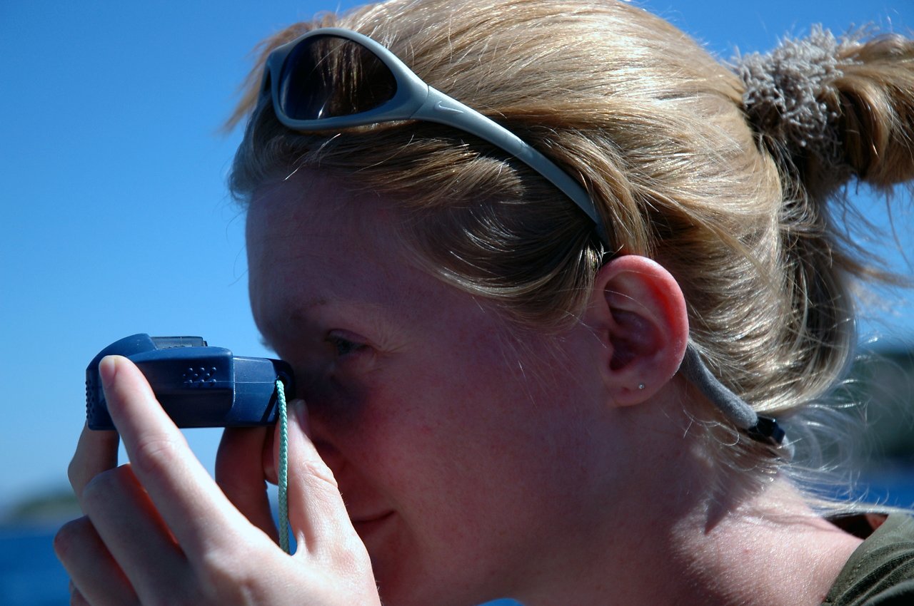 A woman with sunglasses on her head looks through a small blue camera or viewfinder, focusing on something ahead.