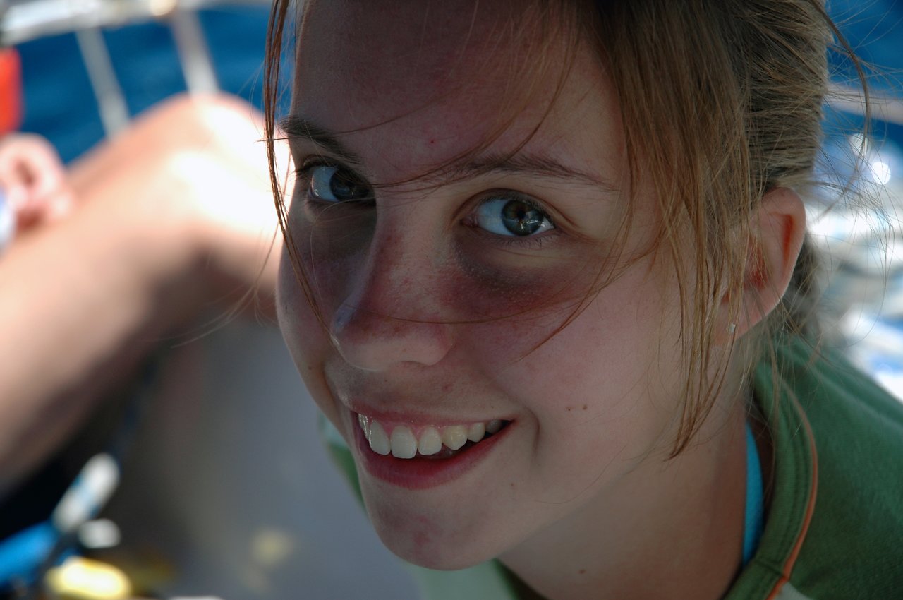 A young woman with light brown hair smiles at the camera, with sunlight on her face.