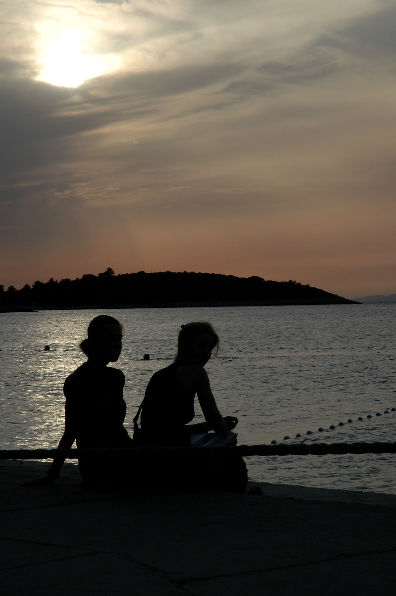 Two people sit by the water at sunset, facing slightly away, with their silhouettes visible against the sky.