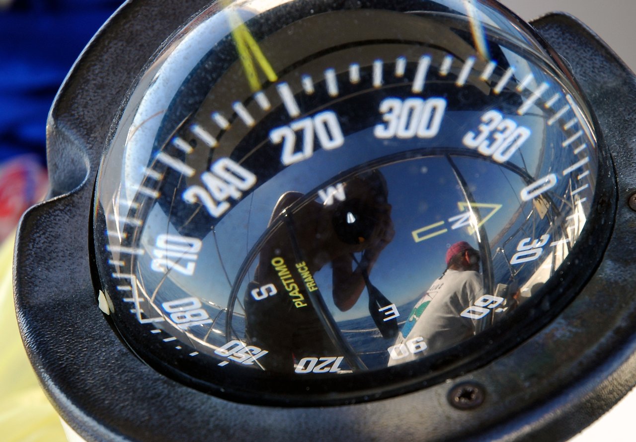 A boat's compass reflects a person taking a photo and another person sitting on the deck, looking at the water.