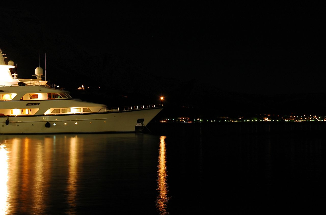 A large yacht with glowing interior lights is docked at night, reflecting on the calm water.