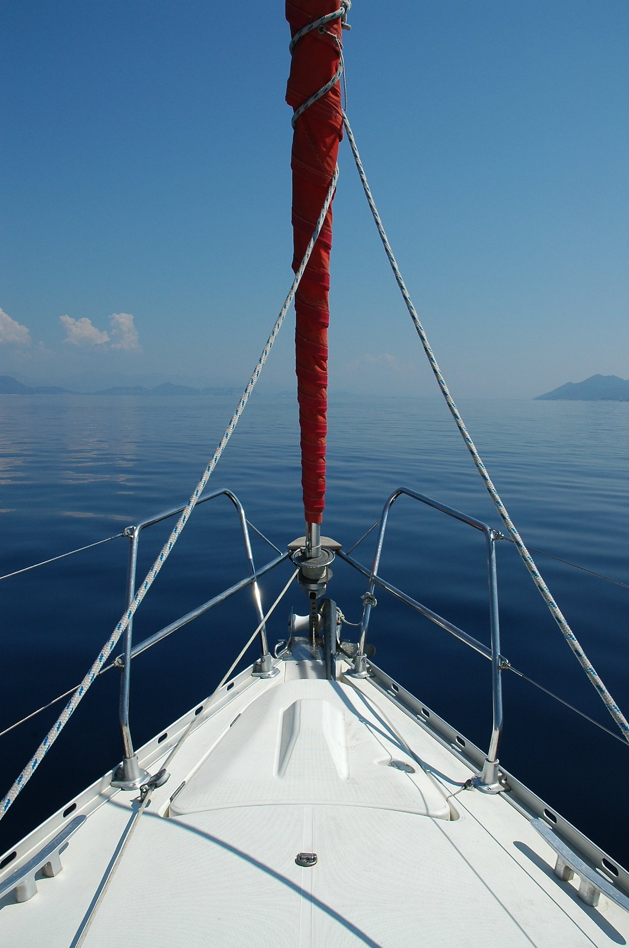 The bow of a sailboat with a furled red sail, floating on calm water under a clear blue sky.