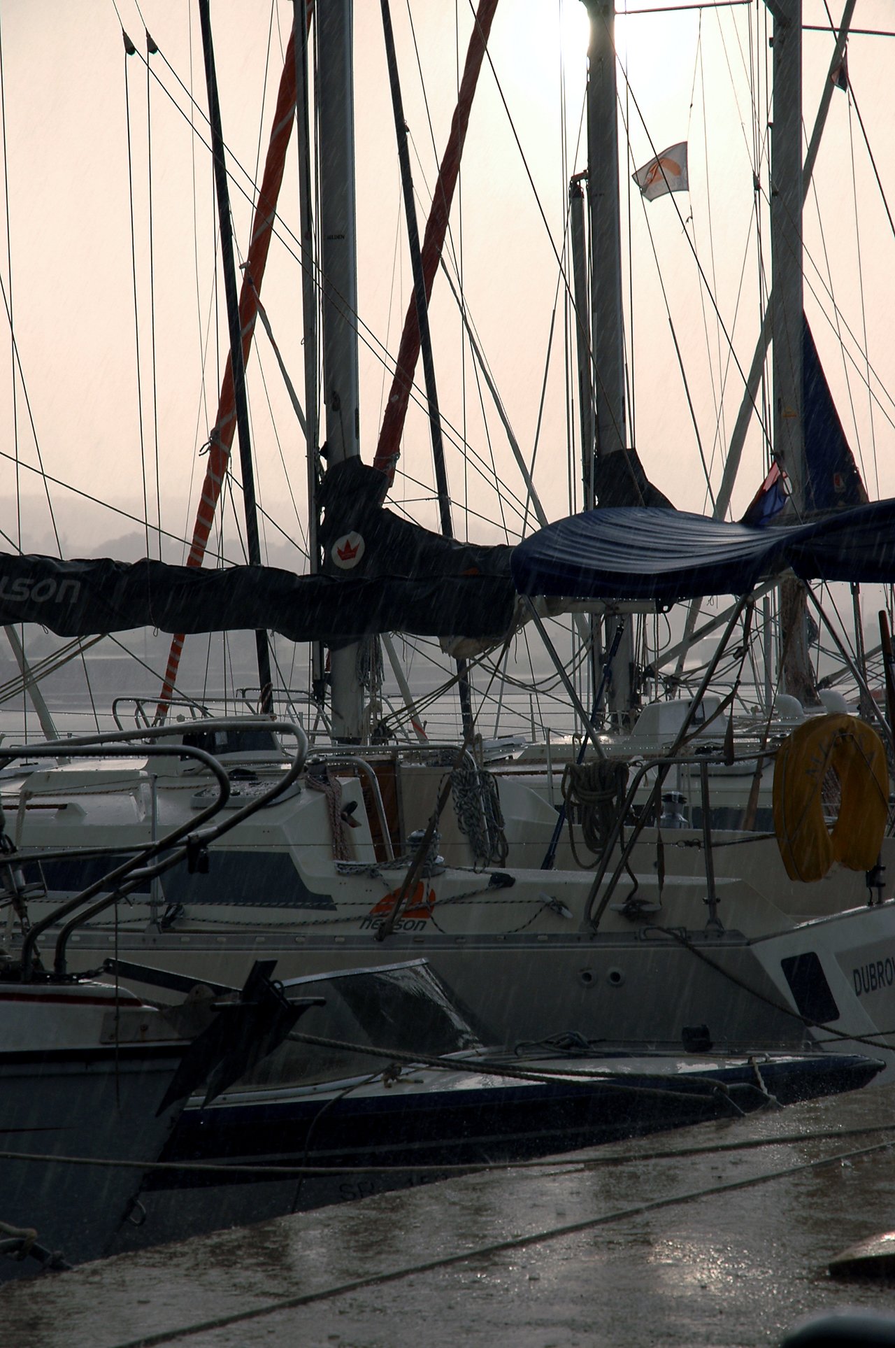 Sailboats docked at a marina in the rain, with wet surfaces and raindrops visible on the boats and water.