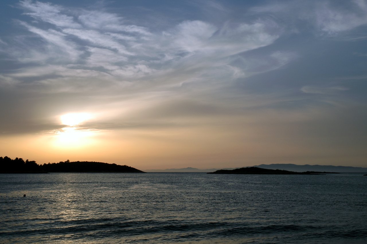 The sun sets over a calm sea, with small islands in the distance and soft clouds in the sky.
