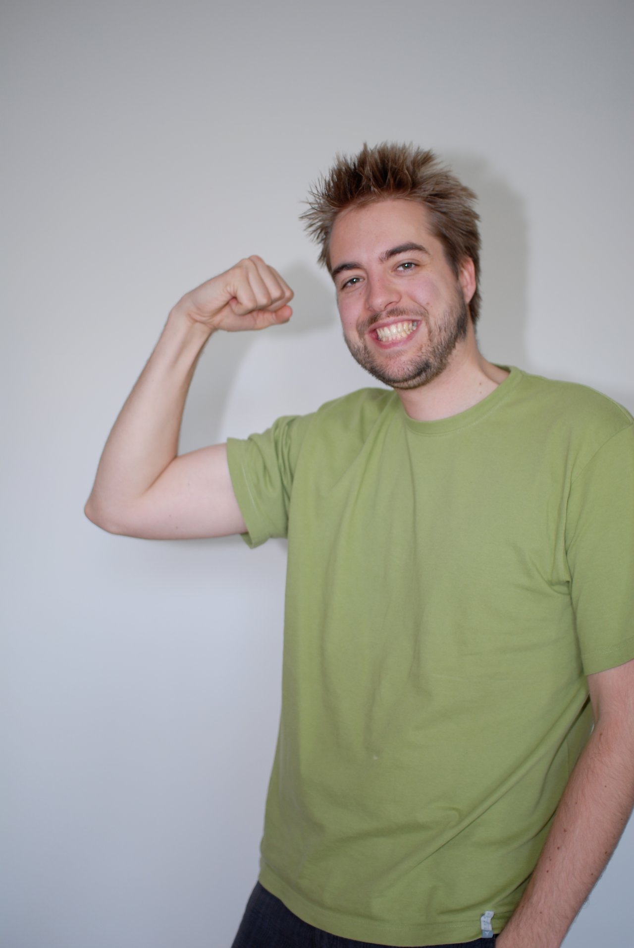A man in a green shirt smiles and flexes his arm in front of a plain background.