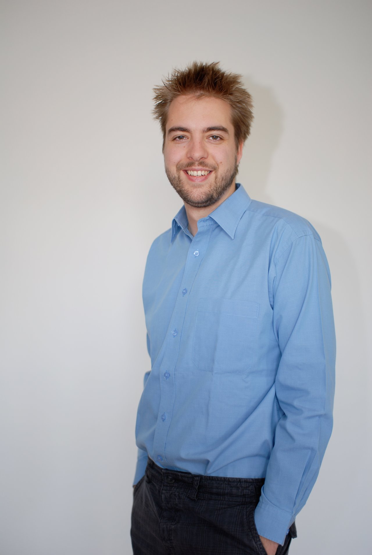 A man with short, spiky hair and a beard smiles while wearing a blue button-up shirt.