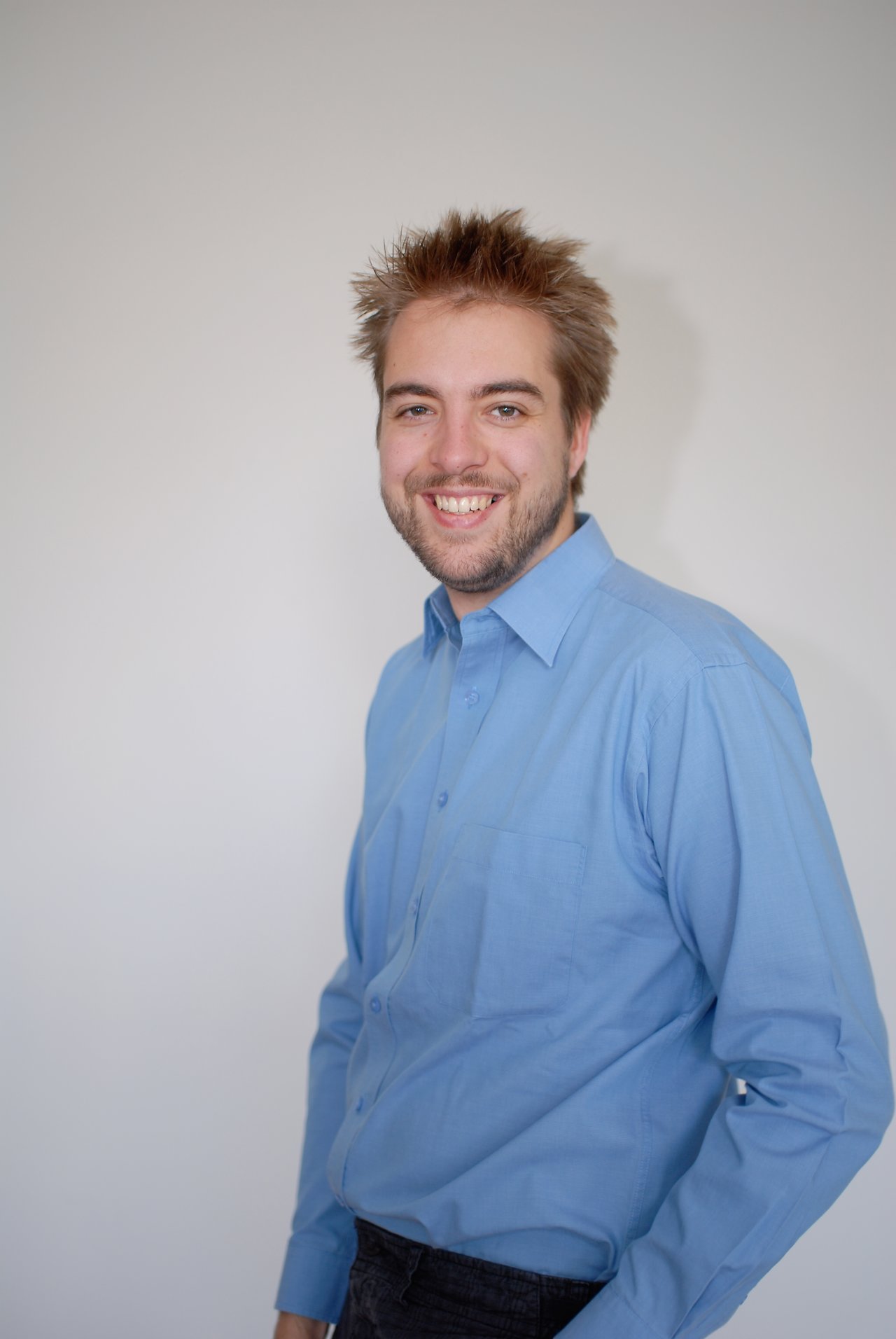 A man wearing a blue shirt smiles at the camera against a plain background.
