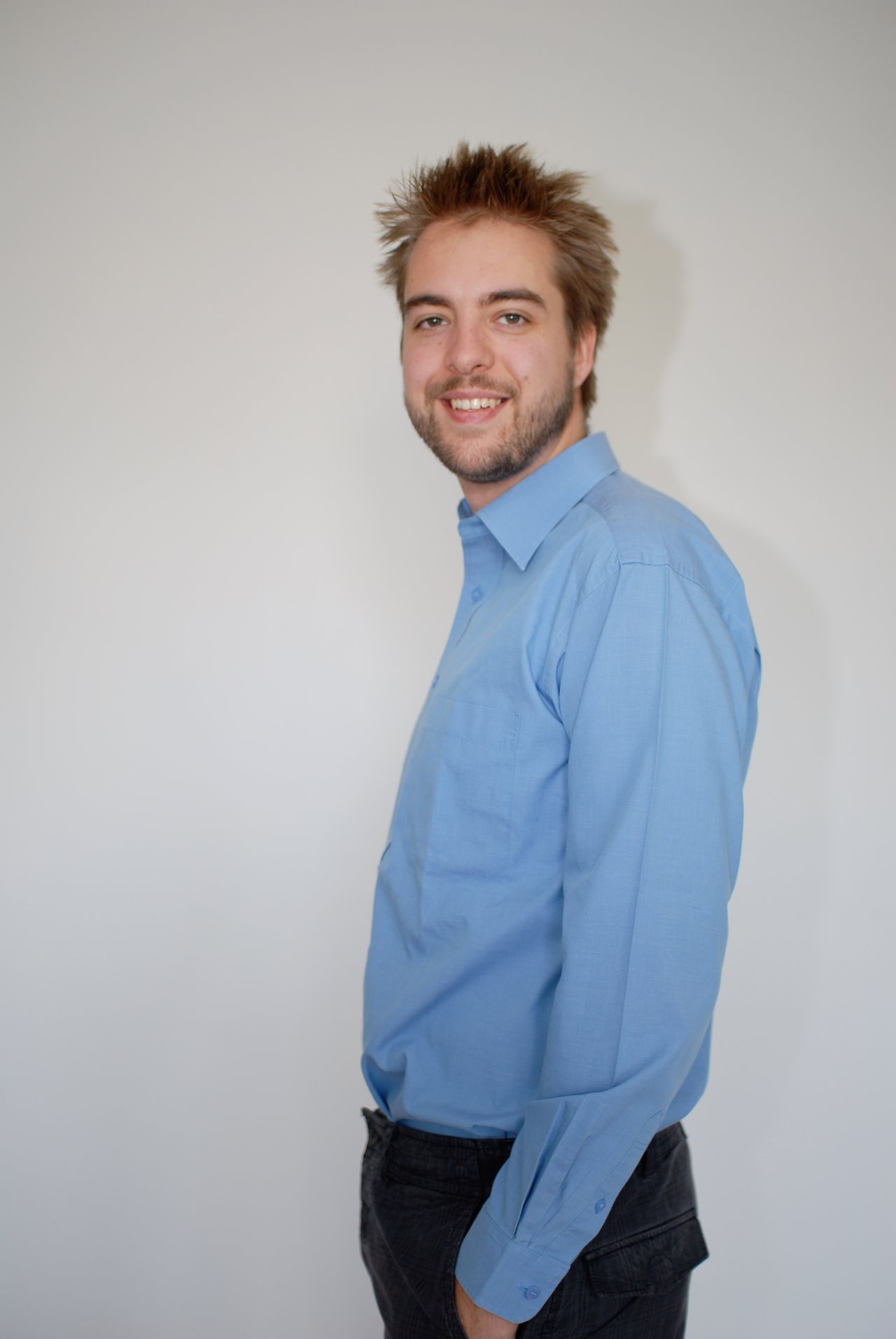 A man wearing a blue shirt stands against a plain background, smiling at the camera.