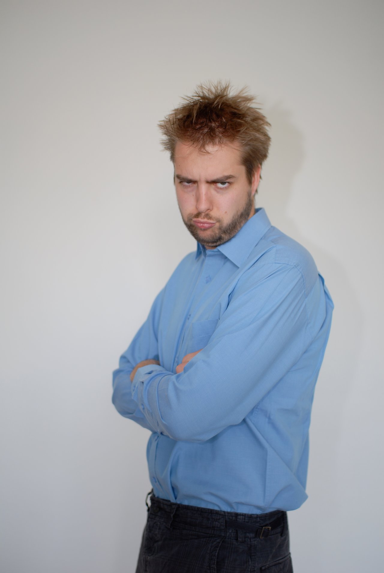 A man in a blue shirt stands with arms crossed, making a playful pout and frowning expression.
