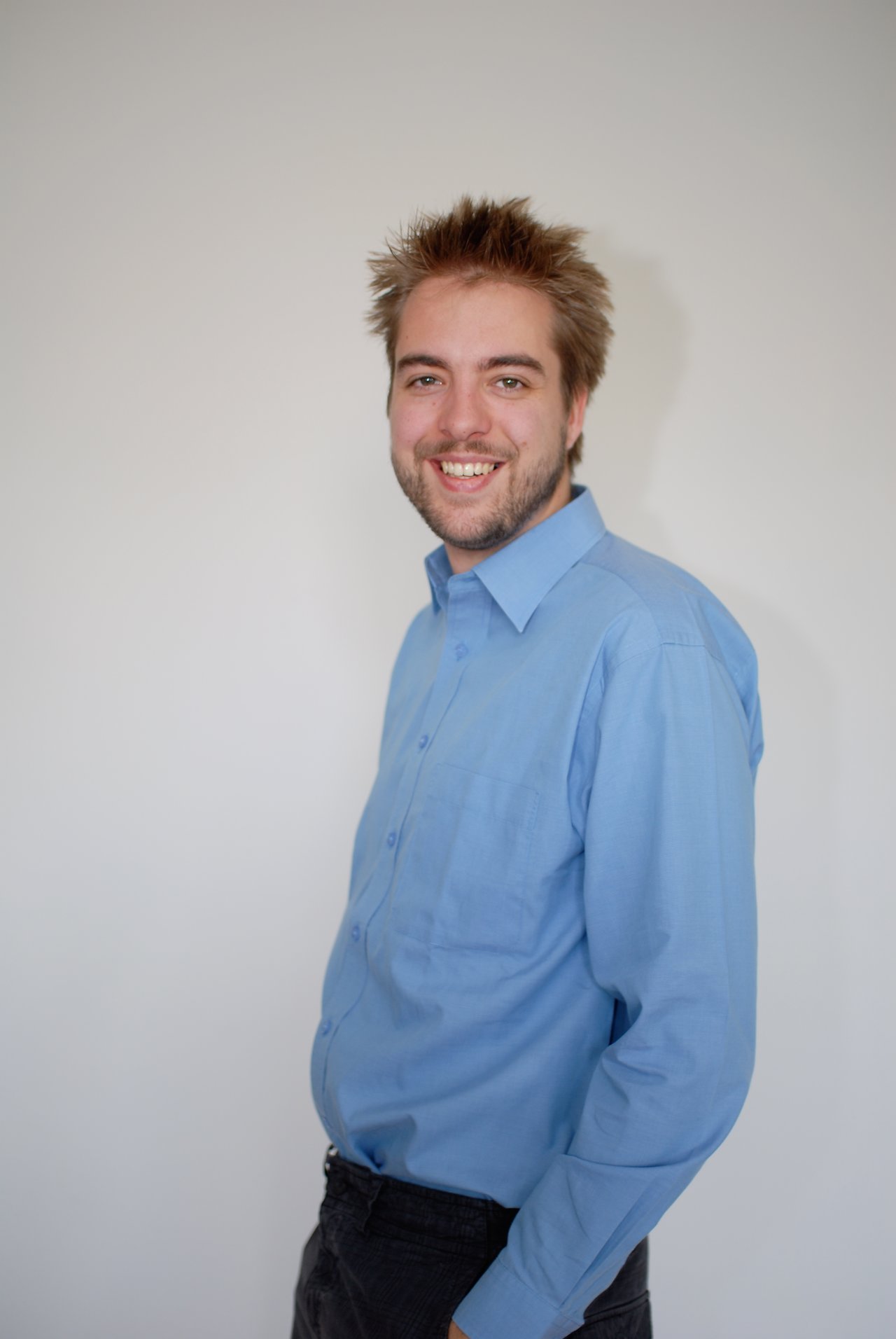 A man wearing a blue shirt smiles at the camera against a plain white background.