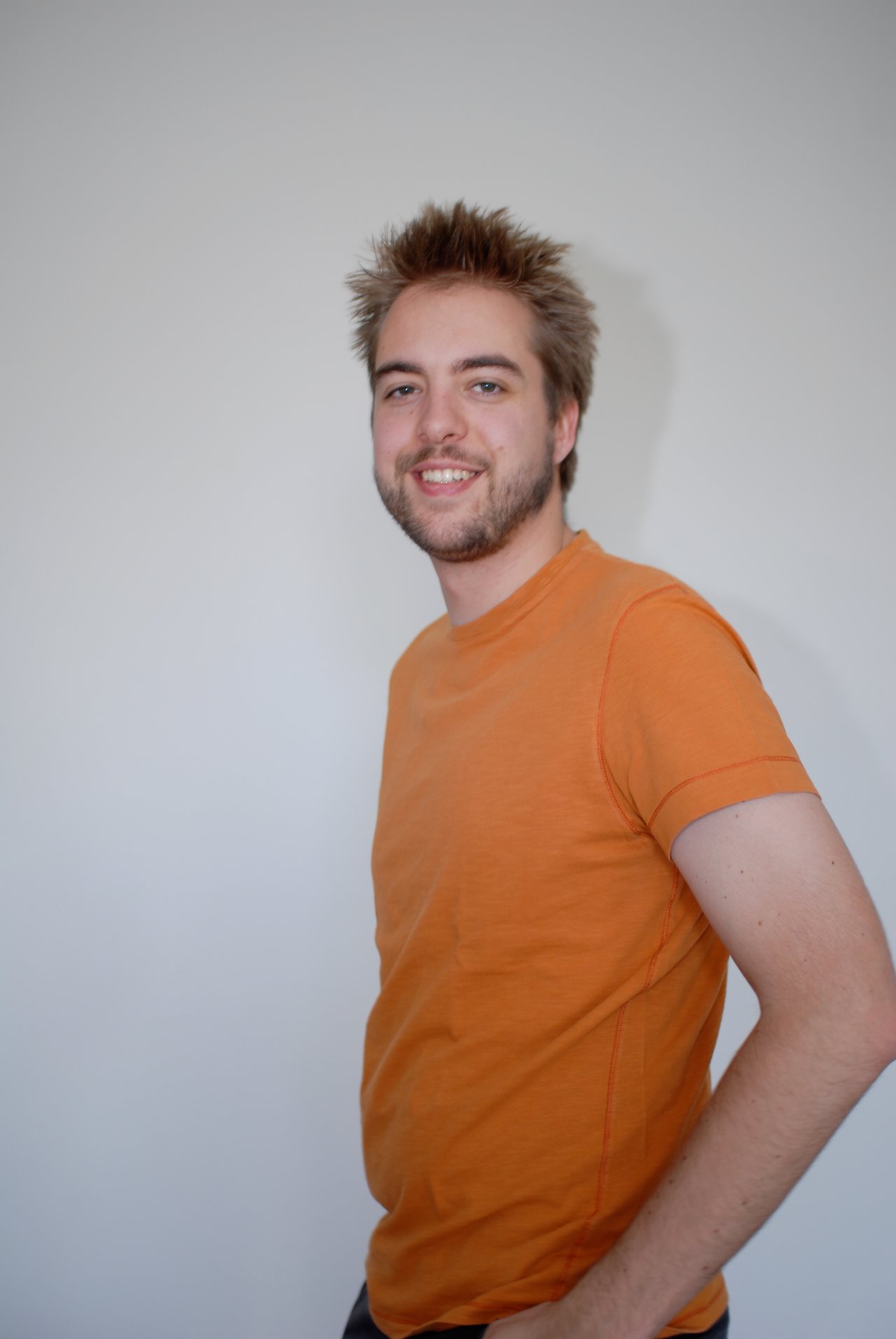 A man wearing an orange T-shirt smiles at the camera while posing against a plain white wall.