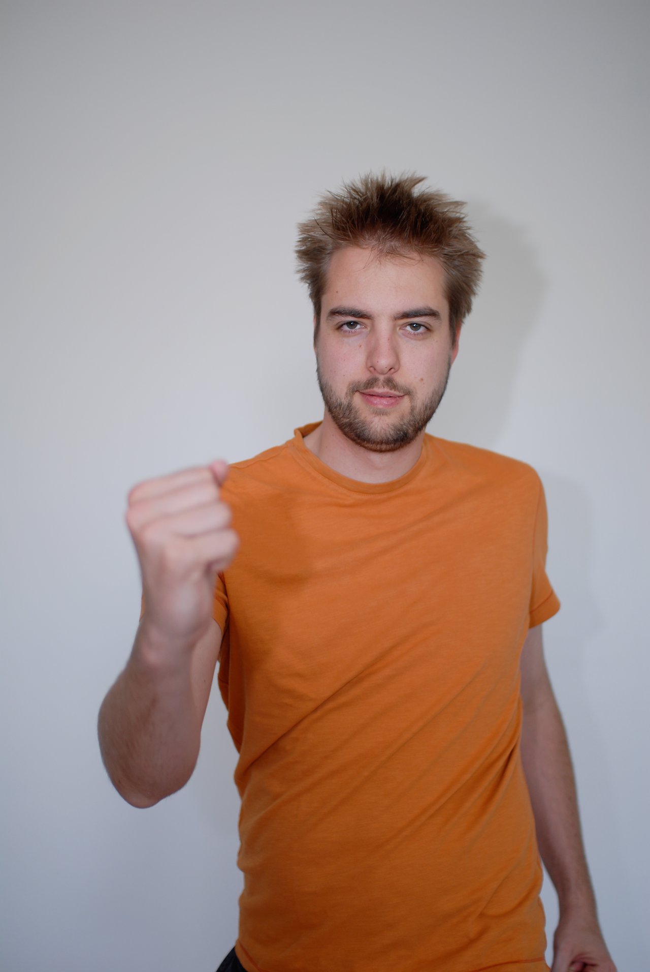 A man in an orange shirt stands against a plain wall, extending his fist toward the camera.