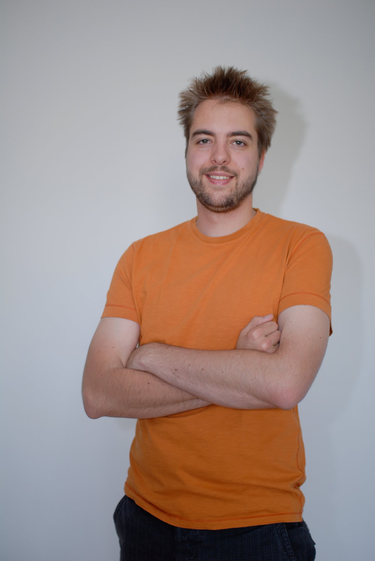 A man with short, spiky hair and a beard smiles with his arms crossed, wearing an orange t-shirt.