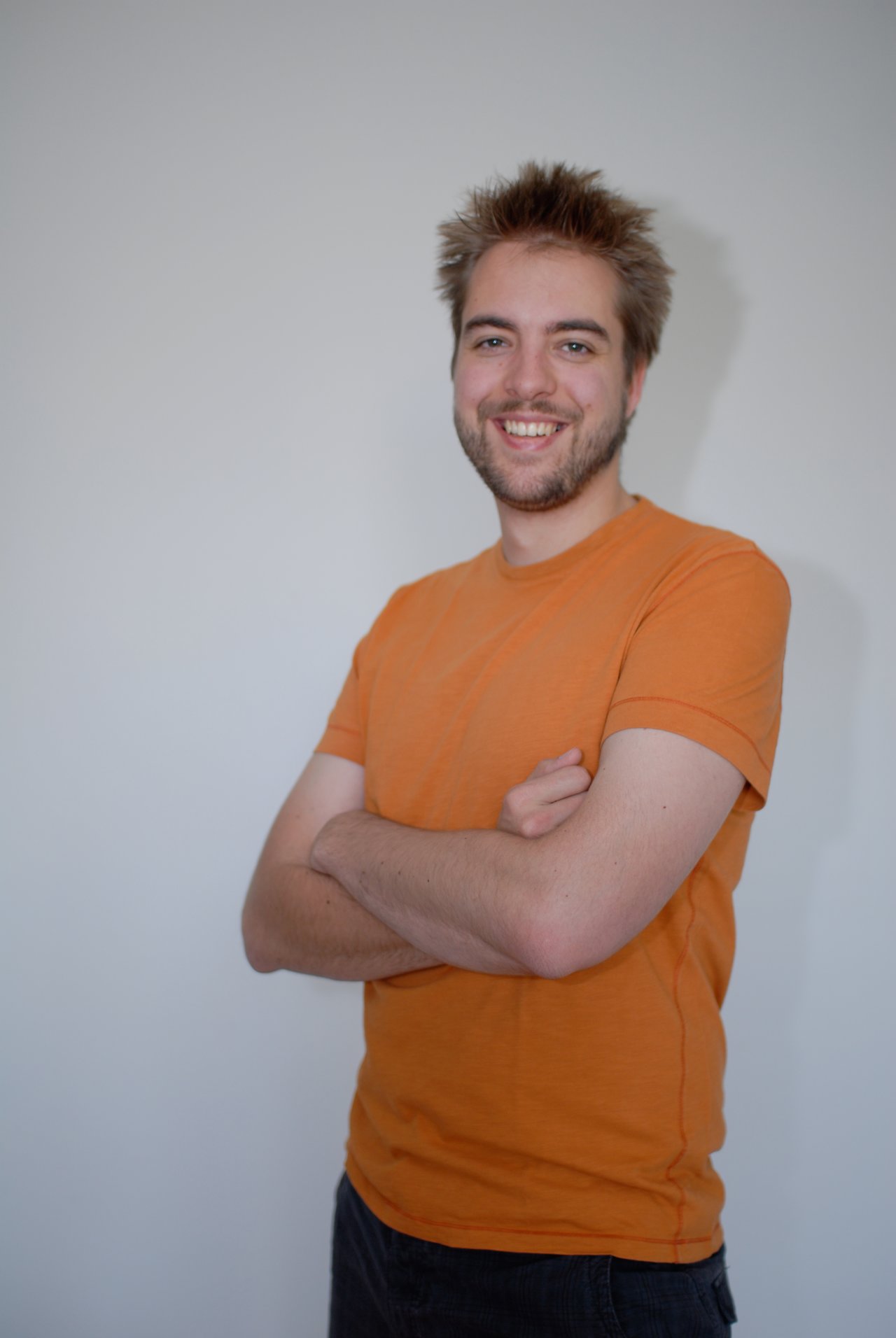 A person wearing an orange shirt stands against a plain background, smiling with arms crossed.