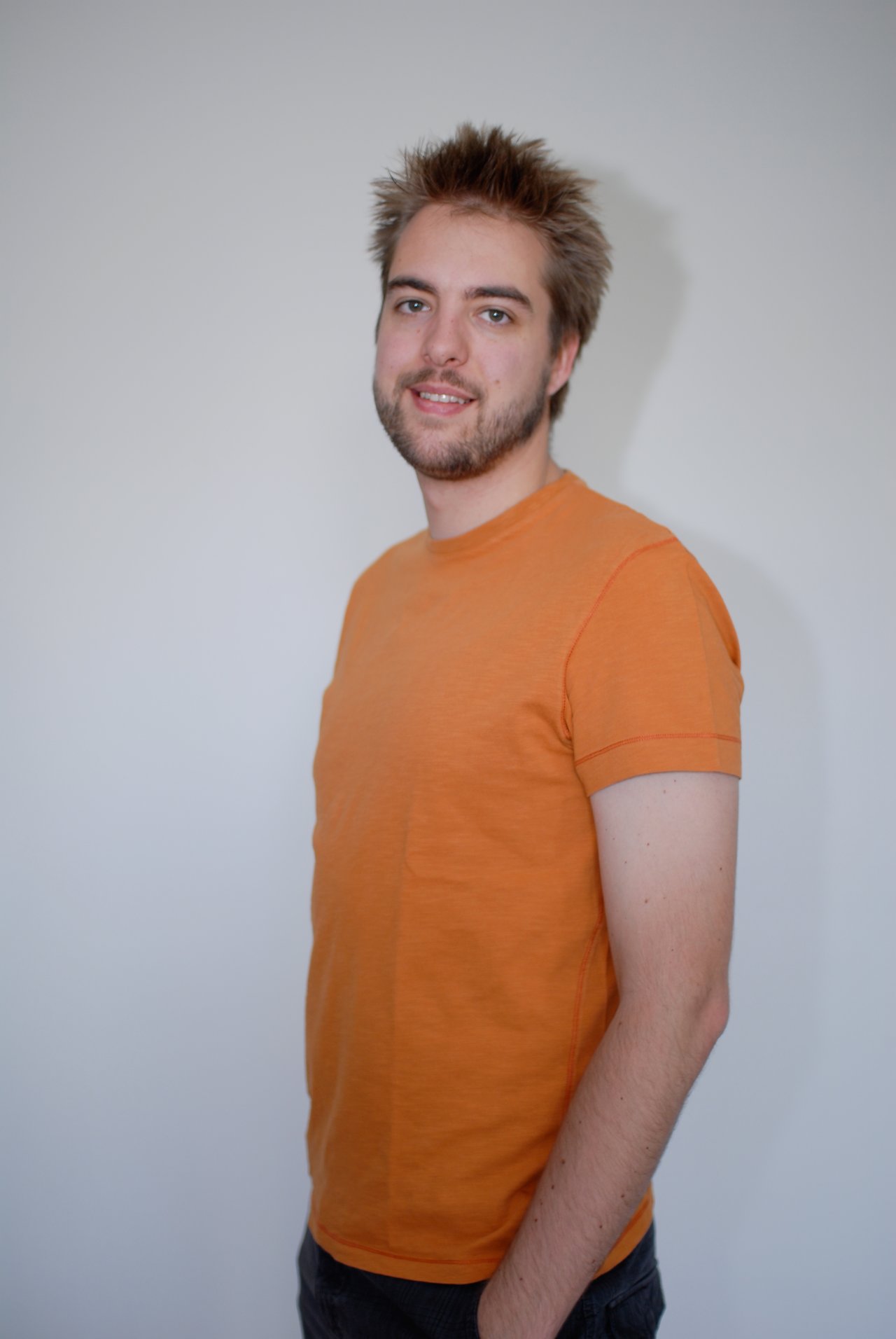 A man wearing an orange T-shirt stands against a plain background, smiling slightly while looking at the camera.