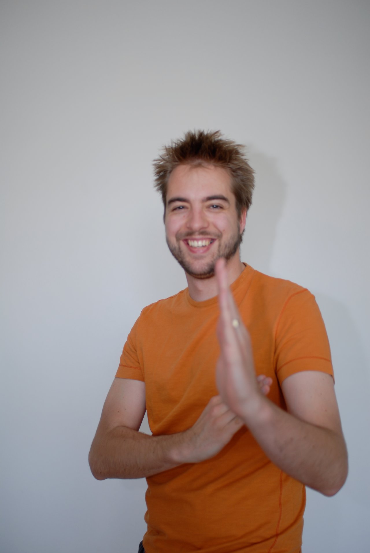A smiling man in an orange shirt poses playfully with one hand raised in front of the camera.