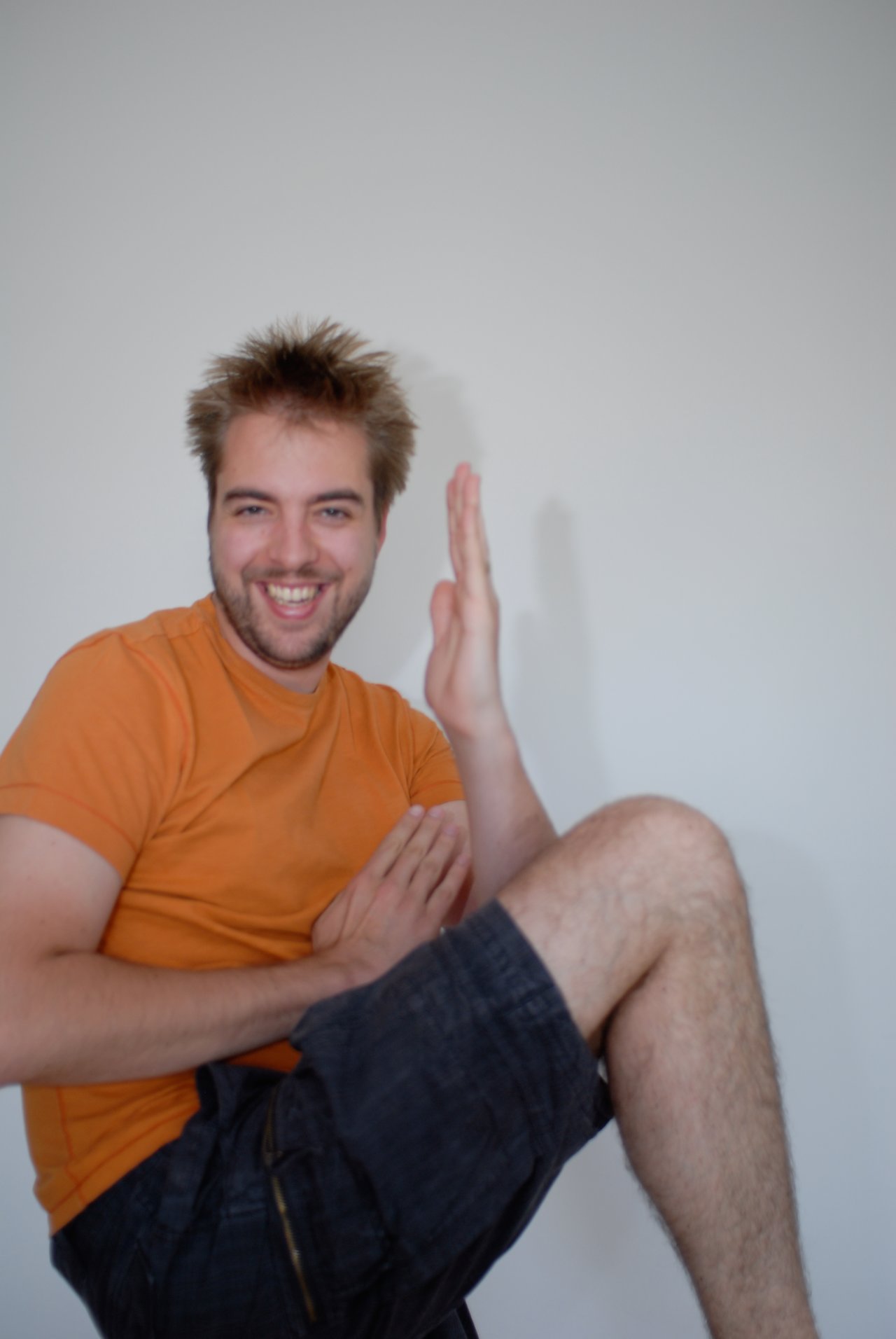 A smiling man in an orange shirt playfully poses with raised hands and a lifted knee against a plain background.