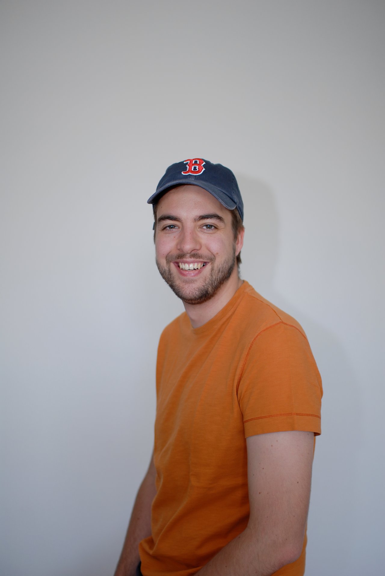 A man wearing an orange shirt and a baseball cap smiles at the camera against a plain background.