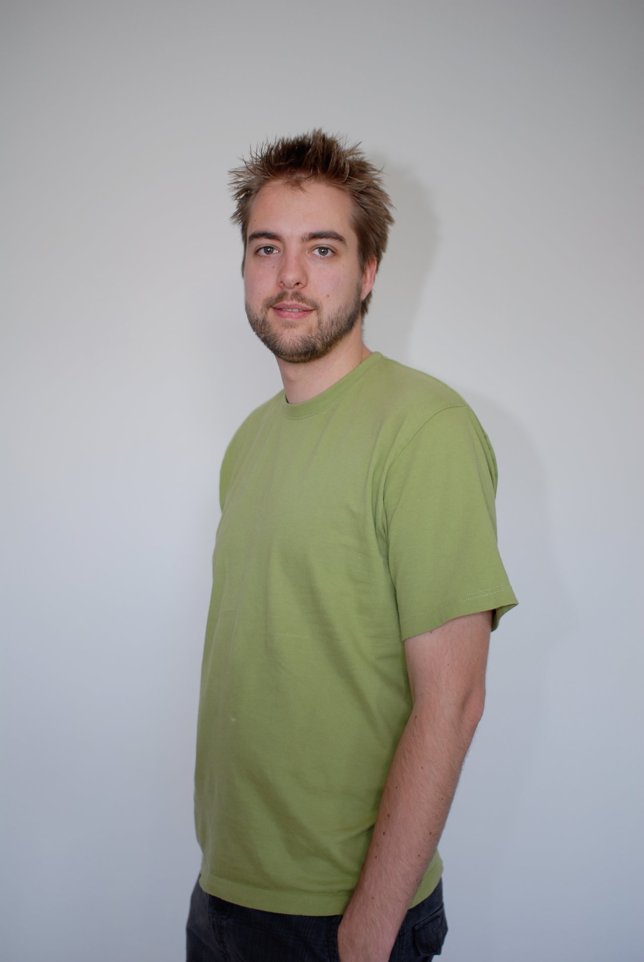 A man with short, spiky hair and a beard stands against a plain wall, wearing a green T-shirt.