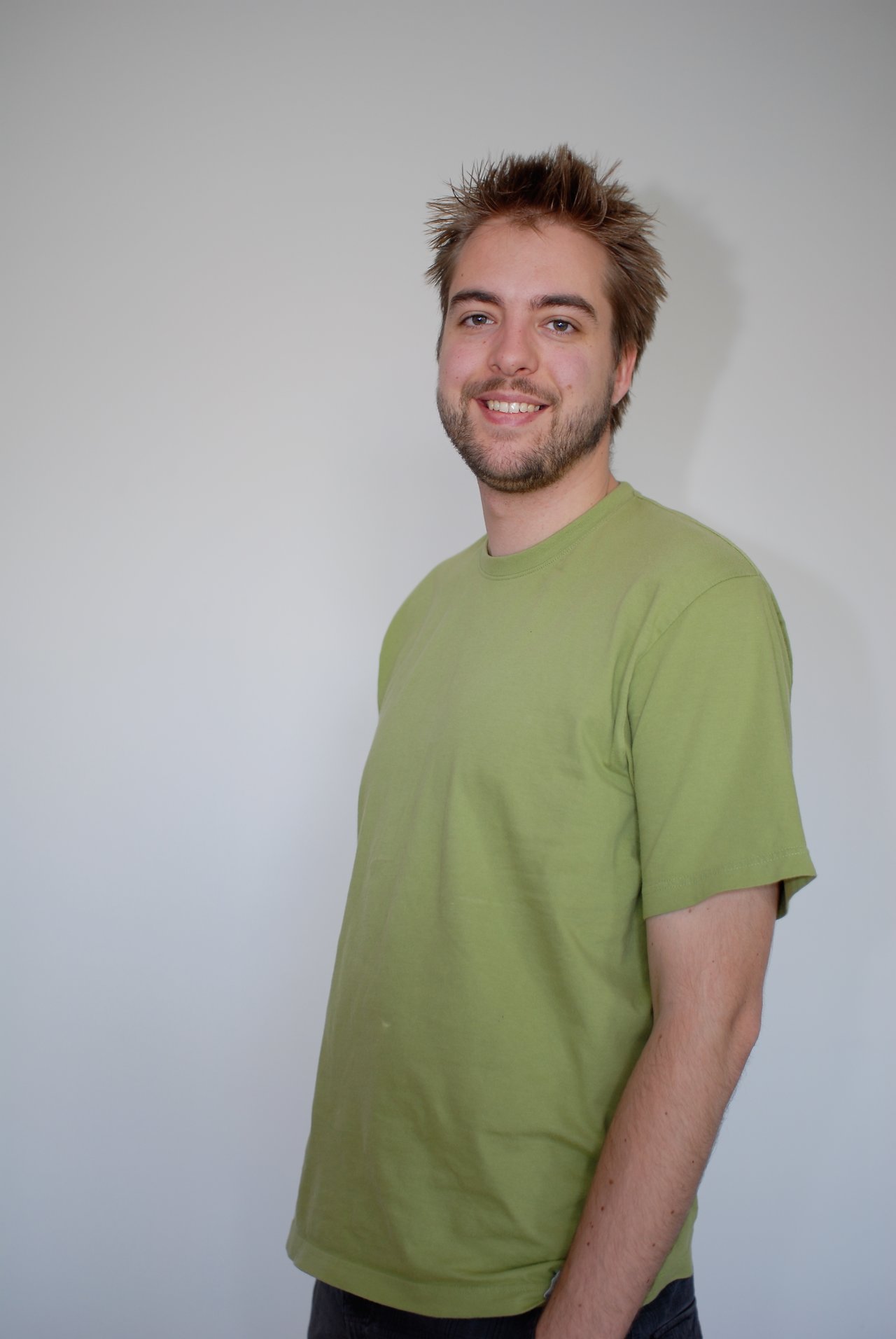 A man with short, spiky hair and a beard smiles while wearing a green T-shirt against a plain background.