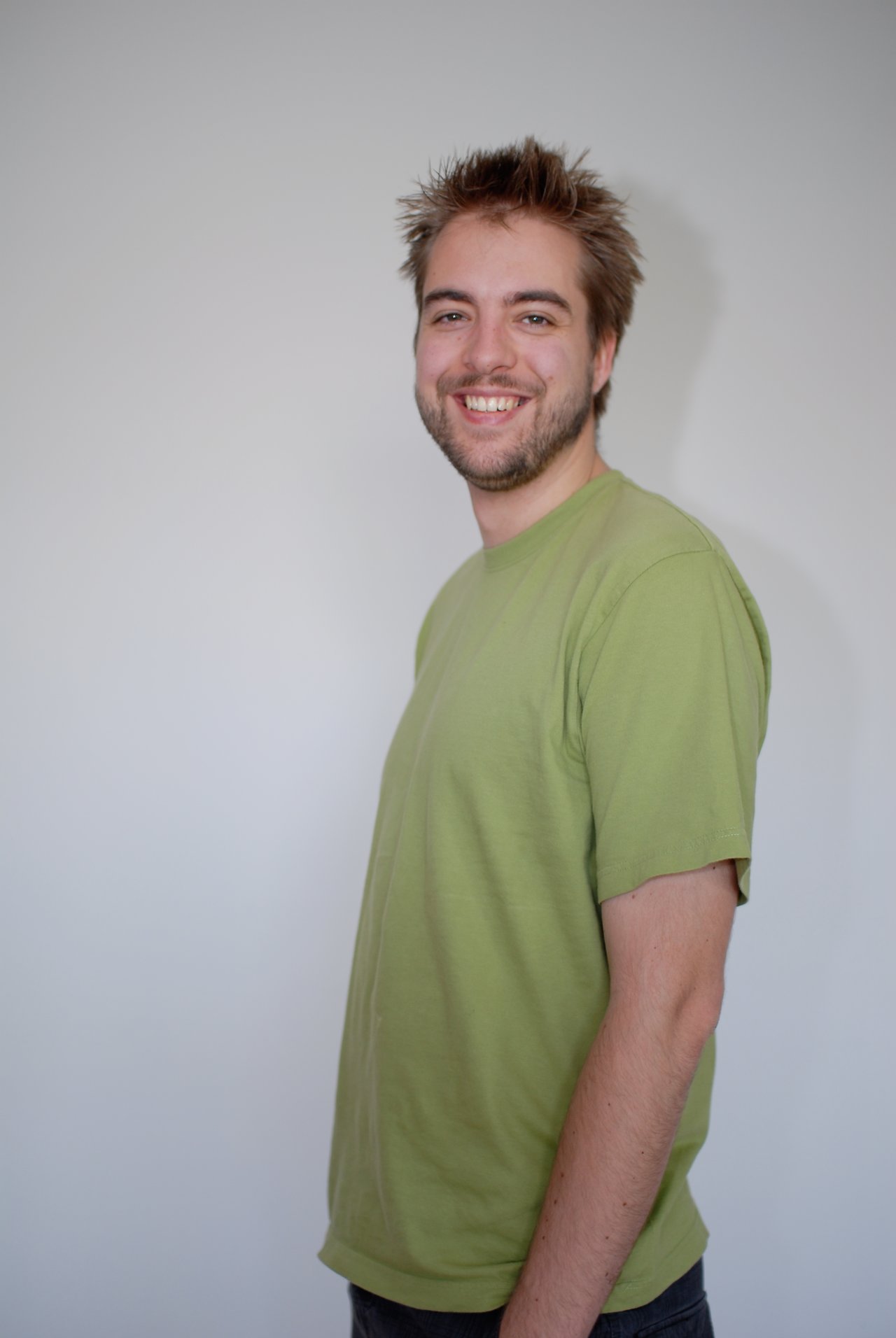 A man with short, spiky hair and a beard smiles while wearing a green T-shirt against a plain background.