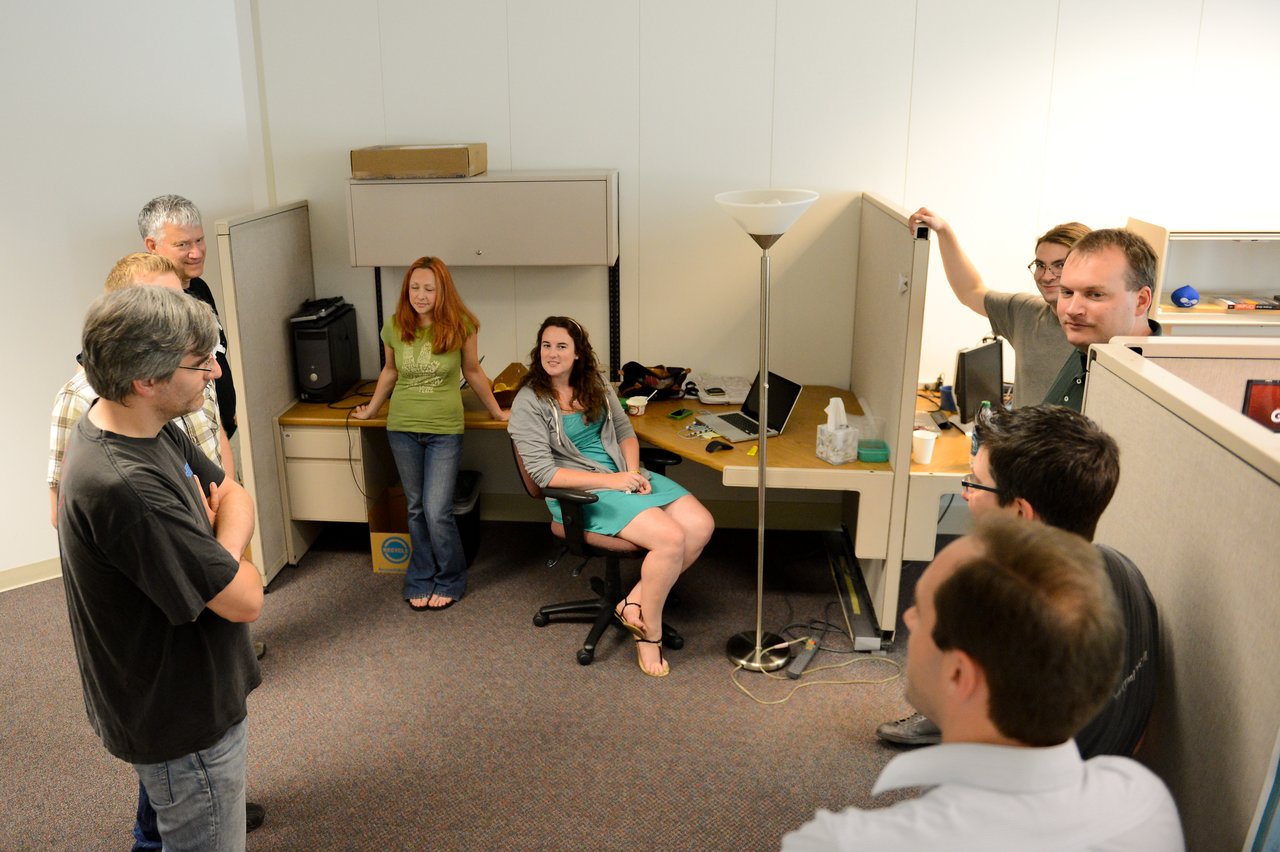 A group of people in an office space are gathered and engaged in a discussion near workstations.