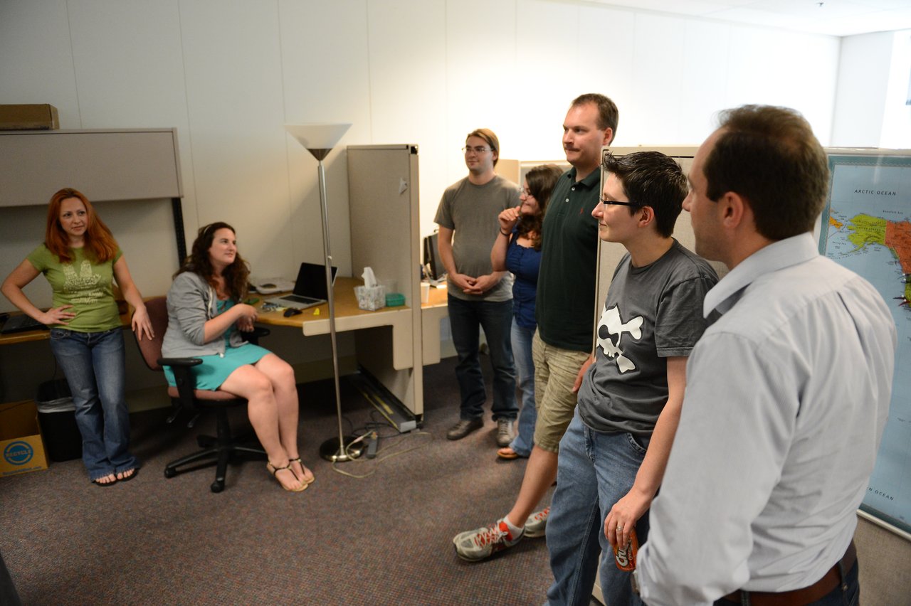 A group of people stands and talks in an office near desks and cubicles, with one person seated.