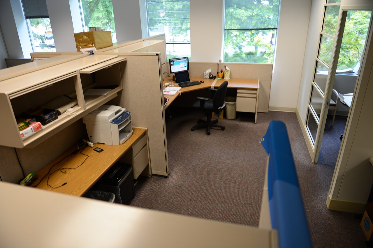 An office workspace with cubicles, desks, a computer, and a printer, featuring large windows and natural light.
